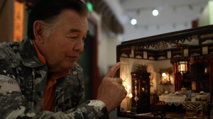 Chinese-American artist Frank Wong peers intently at a detailed diorama that he created of a miniature living room. He points to the inside of the diorama.