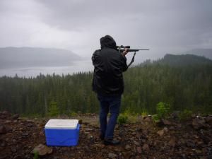 A person wearing jeans and a winter coat pointing a rifle away from the camera. They are in a beautiful natural landscape with trees and water. There is a blue cooler on the rocks next to them.