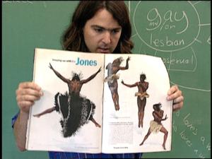A teacher with brown hair stands in front of a chalkboard holding open a book with pictures of acrobatic Black dancers in fabulous costumes. Behind her is a chalkboard with the words gay and/or lesbian inside a circle with lines pointing out of it like a sun.