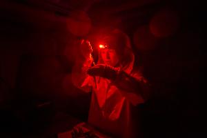 A dark image of a scientist wearing rain gear and a red headlamp looking with curiosity at a vial they are holding.