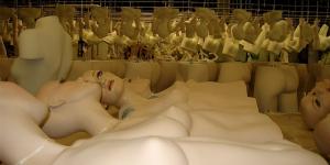 white mannequins lie lined up on a table. Various mannequin parts and metal stands are in the background. The mannequins are bald but have long dark eyelashes and lipstick adorning their vacant expressions.