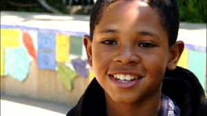 A sweet-faced young Black boy smiling at the camera, in front of a wall with lots of colorful papers attached to it.