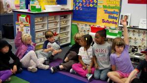 In an elementary school classroom, young students of different genders and ethnicities sit on a purple mat, listening intently, and all looking at someone out of view. One student has their hand raised.