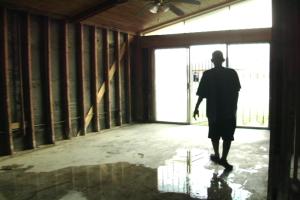 A man stands inside a building where the wall beams are fully exposed and water pools on the cement floor. He is silhouetted against bright windows in the background.