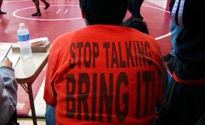 A still from the New Day film Making Noise In Silence. The back of a person sitting on a bleacher. They are wearing a bright orange t-shirt that reads “Stop Talking, Bring It!” in large black letters. The ground is a red gymnasium floor with a wrestling ring.