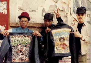 A still from the New Day film My Brooklyn. Two black men stand side by side in front of a white wall with peeling paint. They both wear hats and hold up jackets, the backs of which are decorated with art. The man on the right holds up a peace sign above his head. They are looking straight at the camera with powerful expressions on their faces. Photo by Jamel Shabazz.