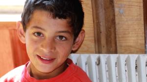 A young Roma boy with tan skin, short dark hair, warm brown eyes and big ears smiles at the camera. He has a smudge on his cheek.