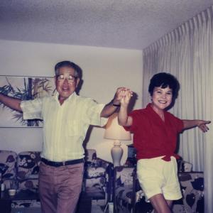 A middle-aged Japanese-American couple hold hands and dance in their living room. Their faces are lit with smiles as they face the camera, arms extended.