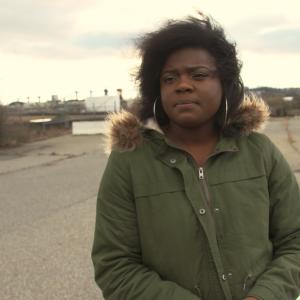 A Black woman looks slightly off camera with a slightly furrowed brow. She wears a winter coat with a fake-fur collar.