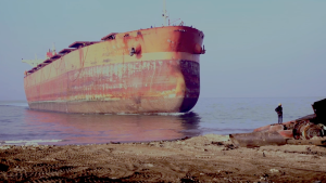 A giant old orange and brown ship sits just a few feet from shore in shallow water. In the foreground a person stands on a pile of junk on the edge of the beach. They are tiny compared to the ship. 