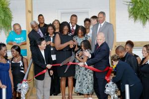 A group of mostly African-Americans of various ages gather on a stairway outside a building while two people hold scissors and are about to cut a red ribbon.