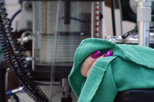 A still from the New Day film Trinidad. A close up of two bright purple nails holding a bright teal cloth. Factory machines fill the blurred background.