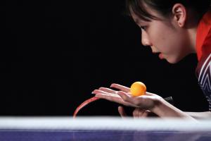 A still from the New Day film Top Spin. An orange table tennis ball rests in Teenage table tennis player Lily Zhang’s open palm. She crouches and looks in the direction of the ball, mouth slightly open in concentration. Her table tennis paddle is visible in her other hand as she prepares to serve the ball.