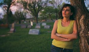 A still from the New Day film Uranium Drive-In. A young woman in a bright yellow sleeveless top crosses her arms and leans against a tree as she looks out thoughtfully into the distance. Behind her are a smattering of graves on bright green grass and more trees.