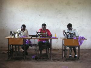 Three Black African teenagers sitting in a row, each at their own foot-pedal operated sewing machine. They focus intently on their work, and two wear tape measures draped around their shoulders.