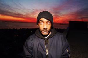 A still from the New Day film When I Came Home. A close up shot of Herold, a war veteran, standing in the center of the frame and staring directly into the camera with a serious expression. He wears a zip-up jacket and a blue beanie. The background is a deep red sunset.