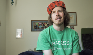 A white man with scruffy beard and a red cap sits in his bedroom laughing. His shirt says Badass Brain Injury Survivor in white screen print.