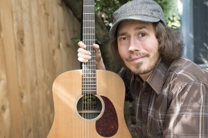 A white man with a scruffy beard and gray cap holds his acoustic guitar and smiles. Half of his face is partially paralyzed.