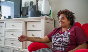 A mixed race woman with short, curly hair sits in an overstuffed bean bag chair in her bedroom smiling.