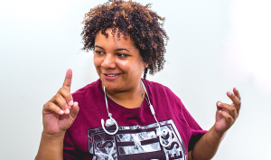 A mixed race woman with short, curly hair smiles while rapping, her hands held up, caught mid-gesture. Earbuds are draped around her neck.
