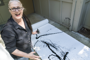 A white woman kneels on her back patio, a huge roll of white paper unrolled by her. She holds a bottle of black ink in one hand and laughs and smiles to the camera. The paper on the ground is decorated with long, arcing lines, splotches, and dots from the ink bottle.