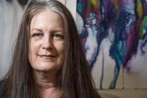 A white woman with long, graying hair smiles softly to the camera. A drippy abstract painting hangs on the wall behind her, jewel tones painted onto white.