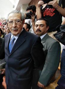 A still from the film “Dictator in the Dock.” An elderly man, General Efrain Rios Montt, and his lawyer are wearing suits and walk, unsmiling, through a crowd of people observing. Several people take pictures of the General as he passes. Another person in a suit stands right behind the General, looking directly into the camera and not smiling.