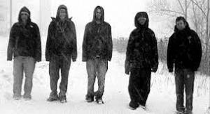 A grainy black and white still from the New Day film Where Soldiers Come From. A row of people shrouded in dark coats and hoods amble down a snowy path. Their surroundings are blurred out with snow but some bushes and trees are visible on the sides of the road.