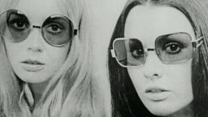 A black and white image from the 1970s of two women, blond and brunette, who stare at the camera, lips parted, with the same still, unsmiling expression. Both wear translucent oversized sunglasses.