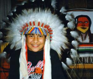 A child with brown skin wears a Native American feather headdress and smiles at the camera. Behind the child on the wall, a blanket and artwork depicting a Native person with long hair.