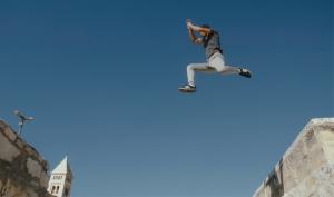 In this low angle shot, the camera is pointed up at a young Palestinian man leaping through the air as he jumps from one building to another.