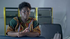 A young Black woman with natural hair sits at a table gesturing as she talks to the camera. She wears a dashiki-style shirt with orange, green and blue patterns. Behind her are three industrial-looking yellow and black shapes.