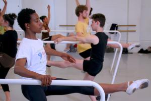 Young ballet dancers of different genders and ethnicities are performing stretches in a dance room. They are holding white bars, and their legs and arms are extended. They wear white ballet shoes.