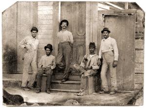 In this old fashioned, grainy, sepia toned photo, three men lean against a doorway, while two sit on a stoop. They wear black hats and stare straight at the camera.