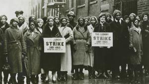 A black and white photo taken in the 1930’s of a group of striking workers who are predominantly Black women. Two women hold signs that say This Laundry is on Strike. Laundry Workers International Union Local 135.