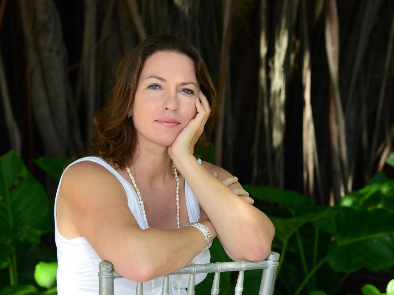 A woman with long brown hair and blue eyes rests on a chair and looks at the camera. 