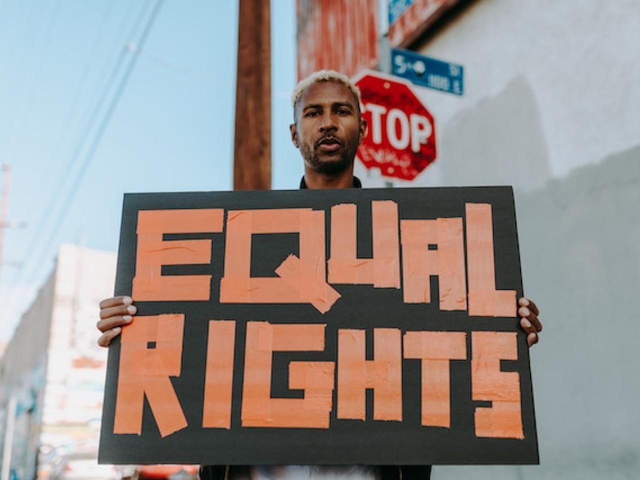 A man is holding up a homemade sign that says "equal rights". There is a stop sign and a cityscape behind him.