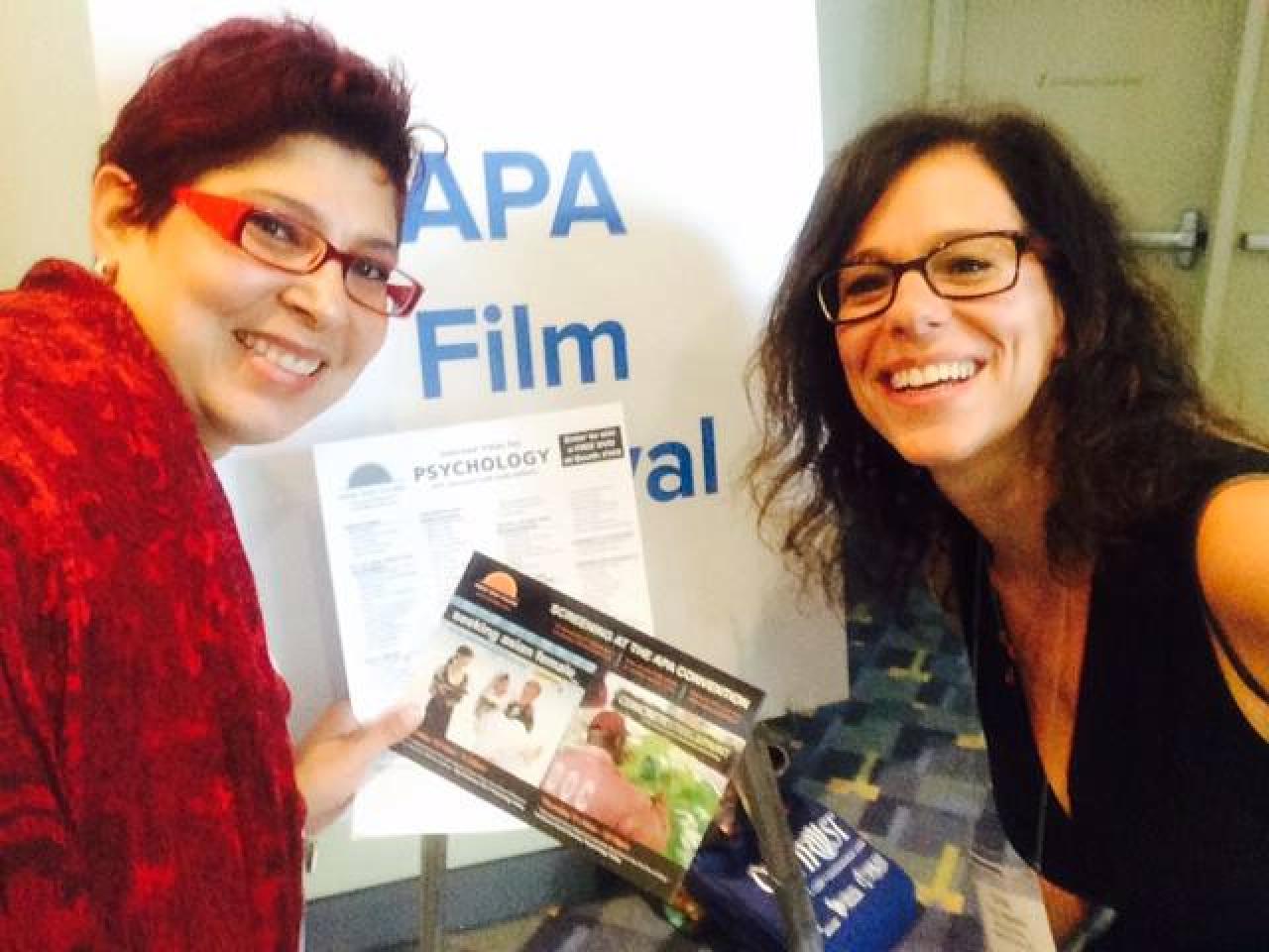 New Day Filmmaker Cindy Burstein and Gigi, from the APA social media, smile and lean into the frame in front of a board that reads “APA Film Festival.” Gigi holds a promotional New Day flyer.