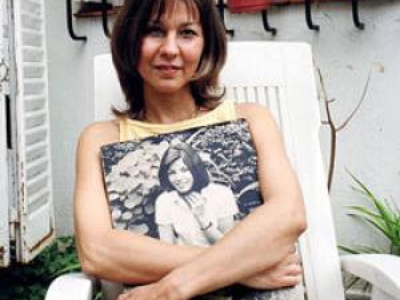 A woman in a sleeveless top sits outdoors on a white lawn chair. She is looking into the camera and her mouth is slightly open in a smile. A black and white image of a smiling young woman rests on her lap, her arms encircling the photograph. In the background are several potted plants and a white window shutter.