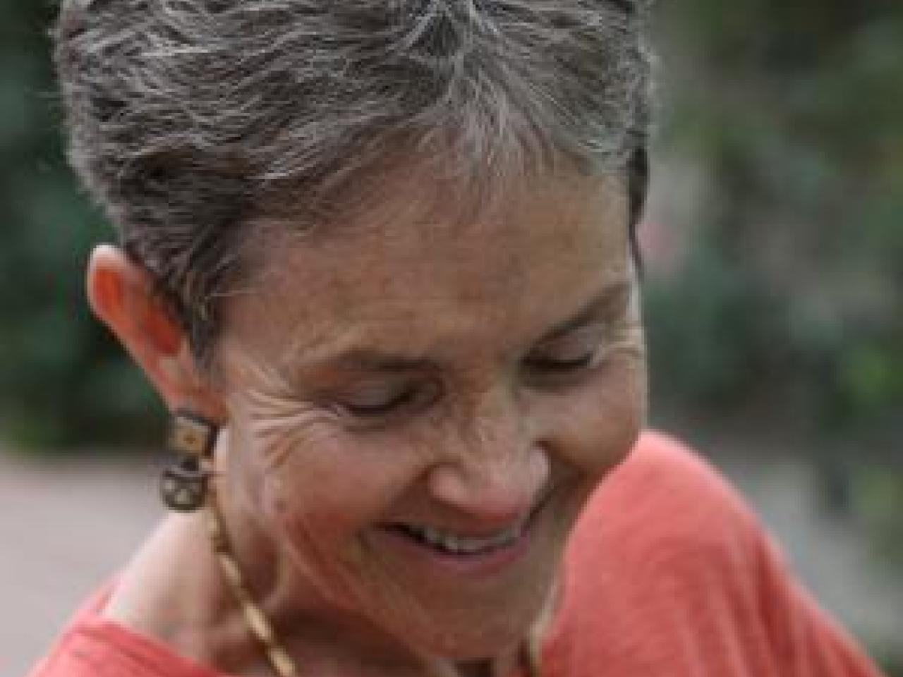 An image from the New Day film States of Grace. Dr. Grace Dammann closes her eyes and smiles towards the ground, her body tilted slightly. Her gray and white hair is short and she wears earrings, a necklace and a long sleeved shirt. The background is full of blurred out foliage.