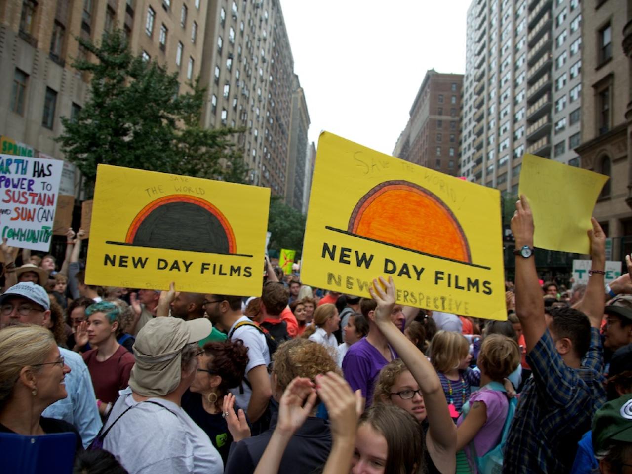 A large group of people of various races, ethnicities and genders mill around in a city street at the People’s Climate March. Several people hold posters up, three of which are yellow films with the New Day Films logo on them. Tall city buildings rise up on either side of the frame.
