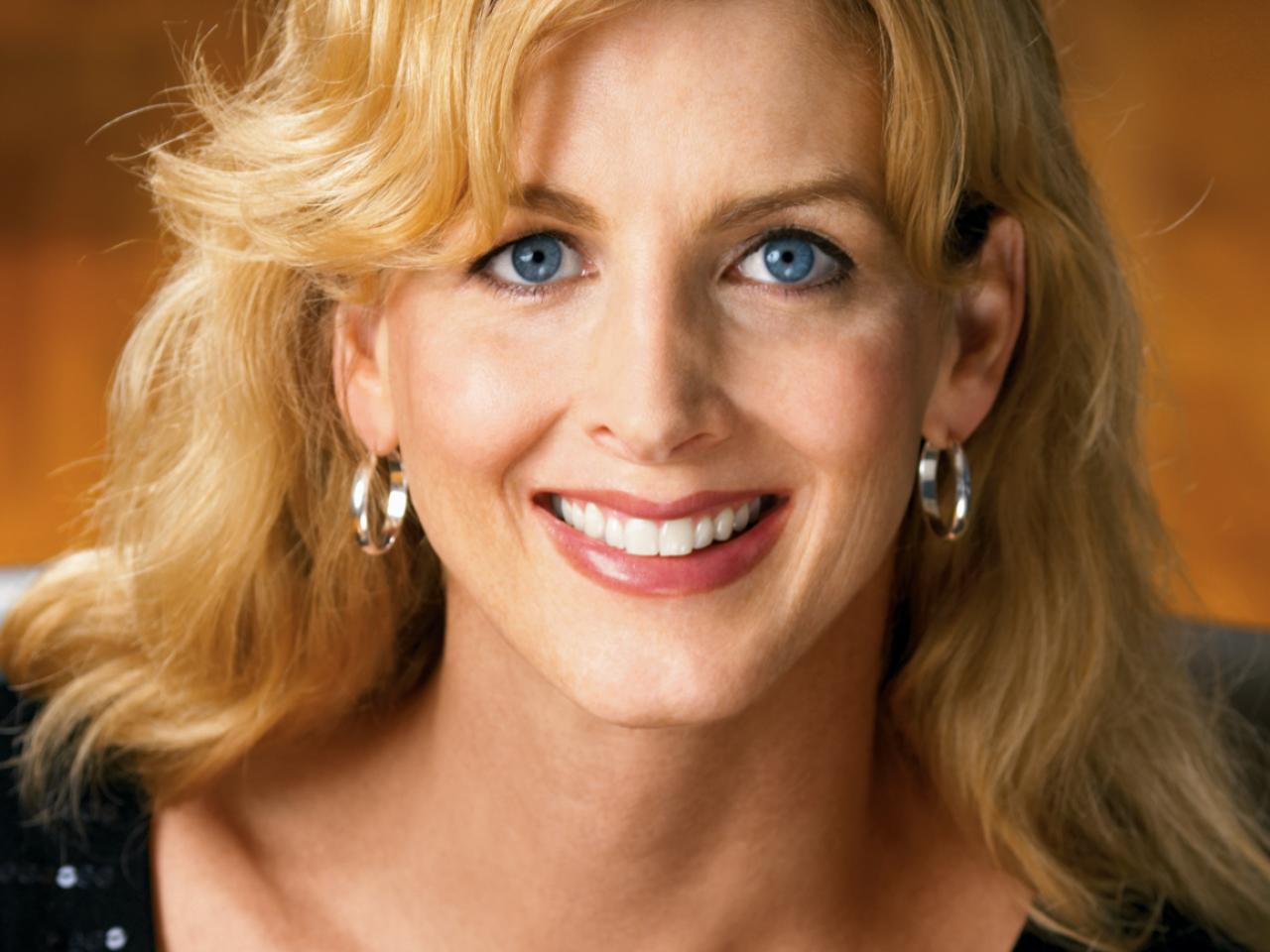 A headshot of New Day filmmaker Kimberly Reed. A trans woman with blonde hair and bright blue eyes smiles widely at the camera wearing hoop earrings. There is a plain orange wall blurred in the background.