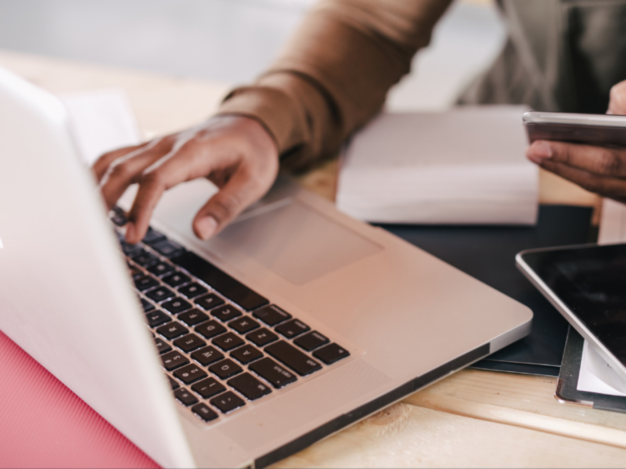 An open laptop on a desk with a hand typing while the other hand is holding a cell phone