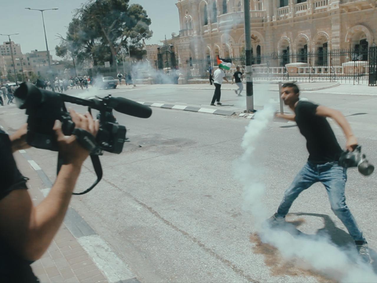 A man holds a tear gas canister and a camera while another films him