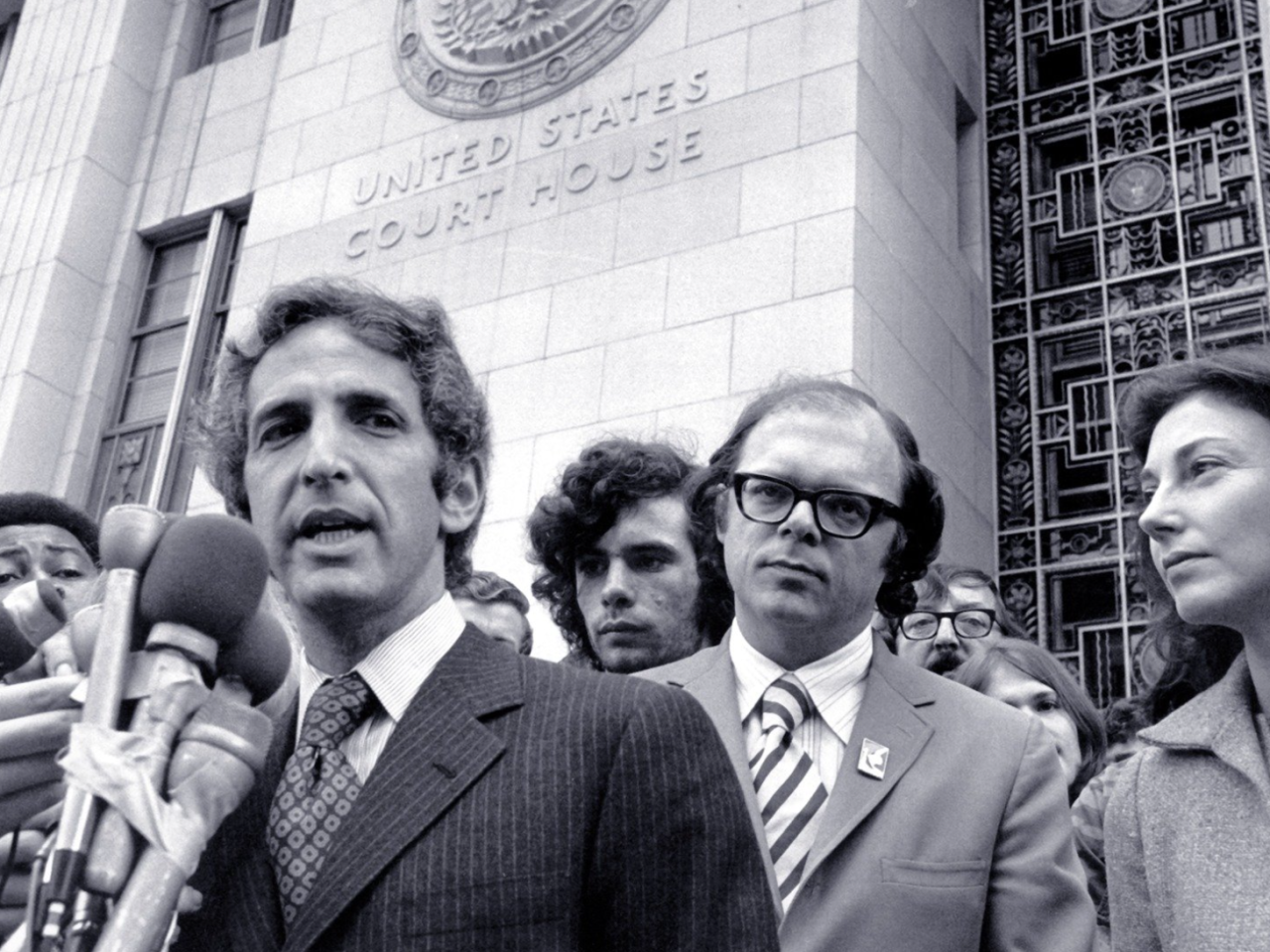 A black and white still image from the film, The Most Dangerous Man in America. The camera looks up from below at Daniel Ellsberg, a white man in a pinstriped suit, who speaks into a mass of TV reporter microphones that are taped together. He stands in front of tall, pale building with “United States Court House” embossed on the exterior wall. He is surrounded by men of various ethnicities who listen on or jockey into position to get their microphone closer to him.