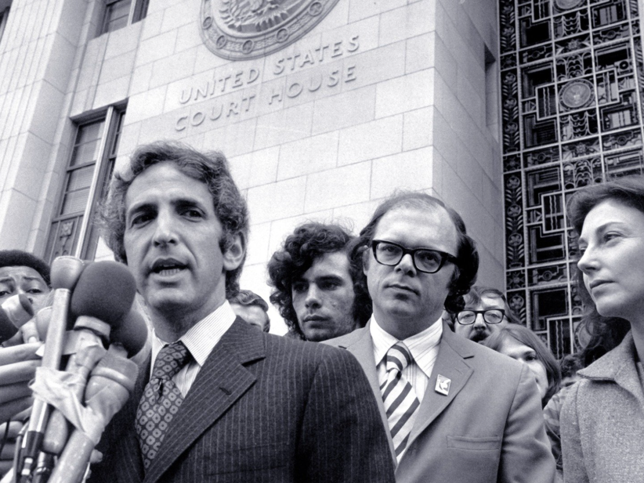 A black and white still image from the film. The camera looks up from below at Daniel Ellsberg, a white man in a pinstriped suit, who speaks into a mass of TV reporter microphones that are taped together. He stands in front of tall, pale building with “United States Court House” embossed on the exterior wall. He is surrounded by men of various ethnicities who listen on or jockey into position to get their microphone closer to him.