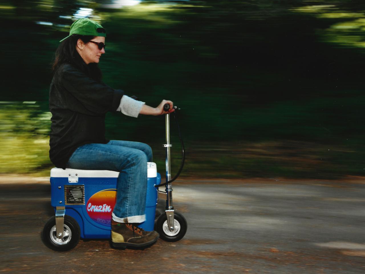 A woman is riding on a vehicle that looks like a cooler with wheels.