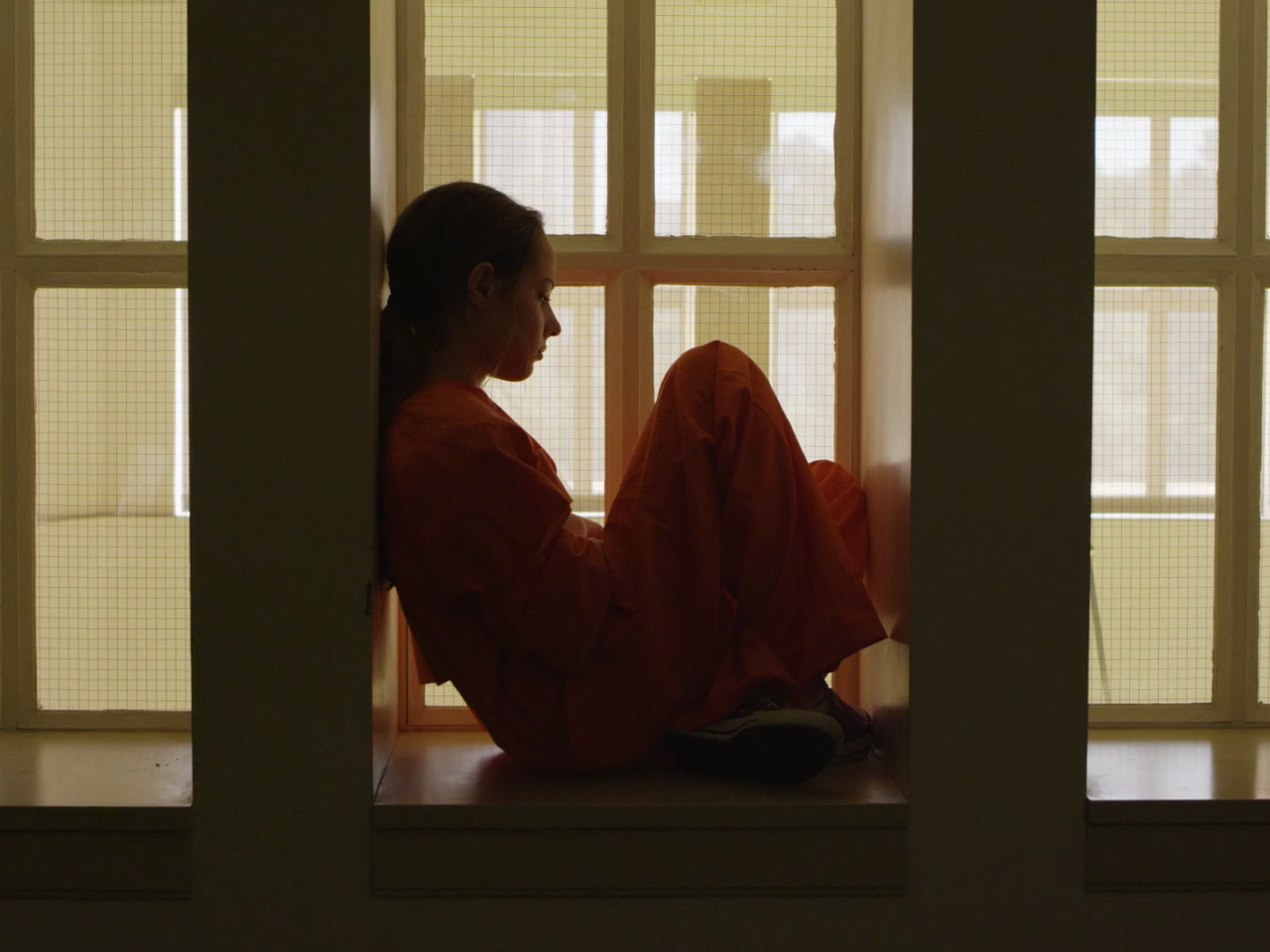 A young woman sits looking out a window in a white room while wearing an orange jumpsuit 