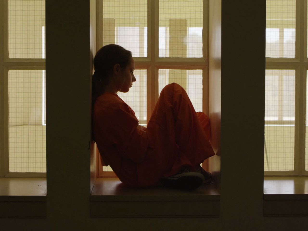 A young woman sits looking out a window in a white room while wearing an orange jumpsuit