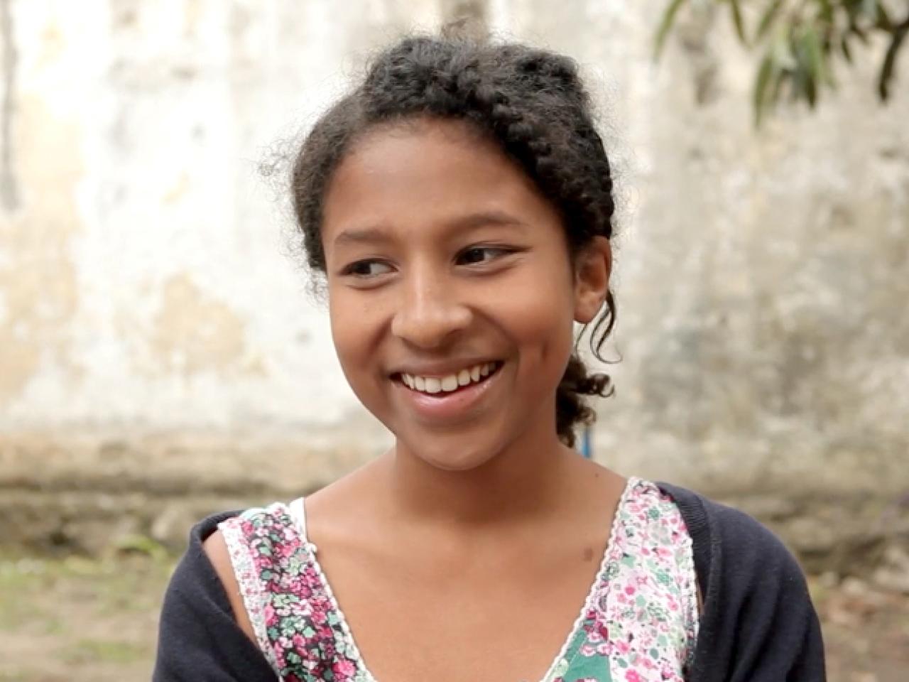 A still from the film The Other Side of the Wall. A girl is standing otside and looks to her left with a slight smile.